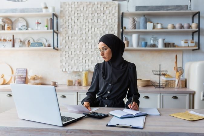 Concentrated young woman in sitting at home at kitchen table and working remotely with documents and laptop.