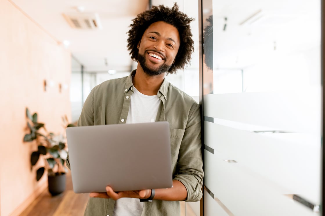 businessman looks at camera and smiles cheerfully, standing in modern office and using laptop.